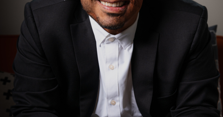 Portrait photograph of a smiling bald Hispanic man seated indoors, wearing a black suit jacket and white shirt. Soft window light and blurred office-like background create a professional, approachable presentation.