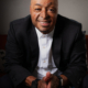 Portrait photograph of a smiling bald Hispanic man seated indoors, wearing a black suit jacket and white shirt. Soft window light and blurred office-like background create a professional, approachable presentation.